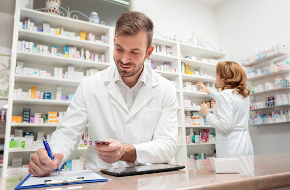 Male-pharmacist-writing-medication-details-and-female-pharmacist-reaching-for-medication-on-shelves